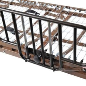 A close-up view of an adjustable hospital bed rail attached to a metal bed frame. The rail is made of dark metal bars and designed to provide safety and support for patients. The bed frame has a grid-like structure for mattress support.