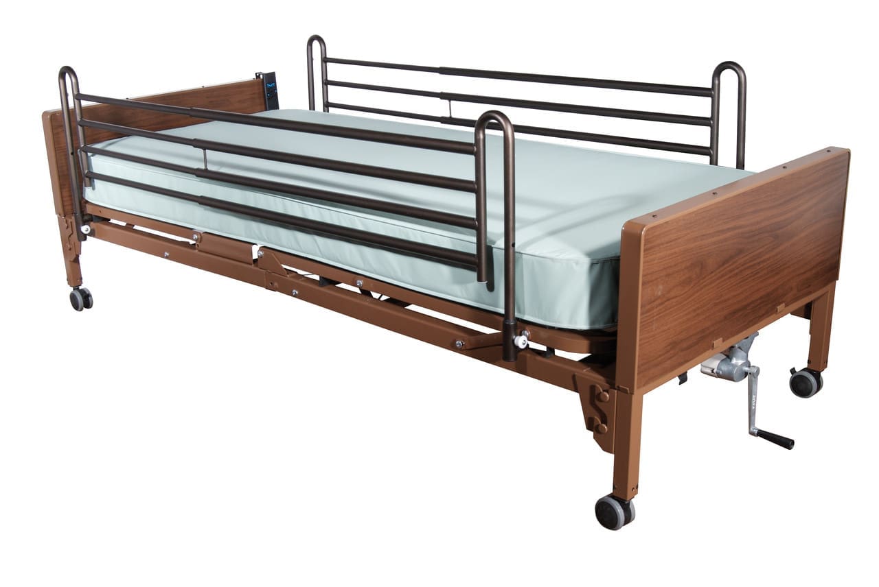 A hospital bed with side rails, a light green mattress, and a wooden headboard and footboard. The bed is on wheels and has a manual crank for adjustments.