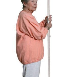 Elderly woman in a peach sweater and white pants stands holding a tall, white support pole, which likely assists with balance and stability.