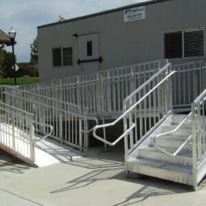 A modular ramp system with railings leads up to a portable building entrance. The ramps ensure accessibility, connecting the building with the surrounding concrete area. A small house and a lamp post are visible in the background.