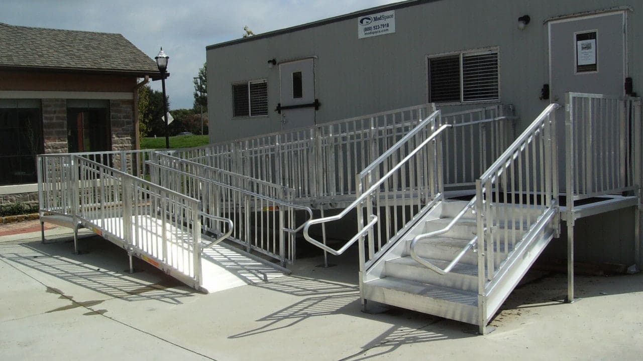 A modular ramp system with railings leads up to a portable building entrance. The ramps ensure accessibility, connecting the building with the surrounding concrete area. A small house and a lamp post are visible in the background.