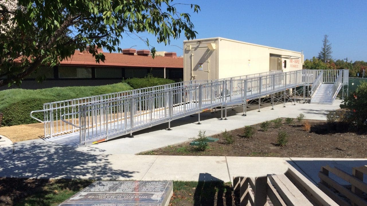 A long silver accessibility ramp leads to a portable building. Surrounded by greenery, the ramp is set on a smooth concrete path with gentle slopes. Trees partially shade the area, and a red-roofed structure is visible in the background.