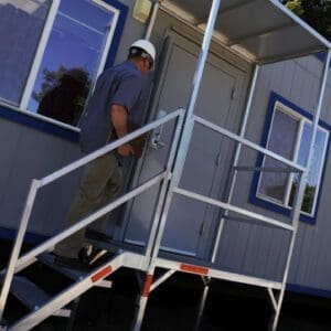 A person wearing a hard hat is ascending metal steps to enter a portable building or trailer, set outdoors with surrounding greenery. The structure has multiple windows and a door with a small canopy above.