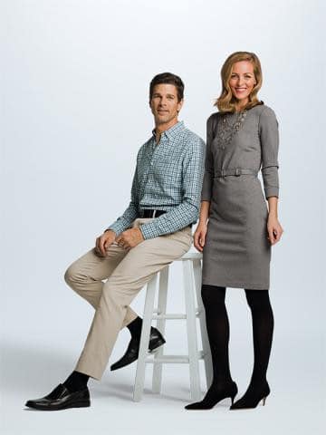 A man and a woman pose against a plain background. The man, seated on a stool, wears a checkered shirt and beige trousers. The woman stands beside him, dressed in a fitted gray dress with black tights and heels. Both are smiling.