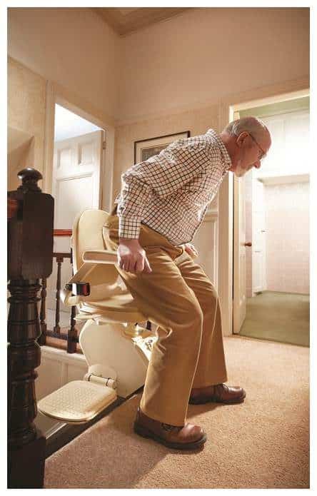 An elderly man is seated on a stair lift at the top of a carpeted staircase in a home. He holds the armrests as the lift prepares to descend. He is wearing a checkered shirt and beige pants.