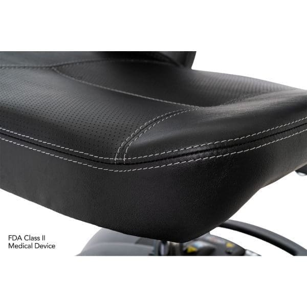 Close-up of a black leather chair with visible white stitching. The chairs perforated surface suggests comfort and durability. A small part of a control panel is visible below. Text reads: FDA Class II Medical Device.