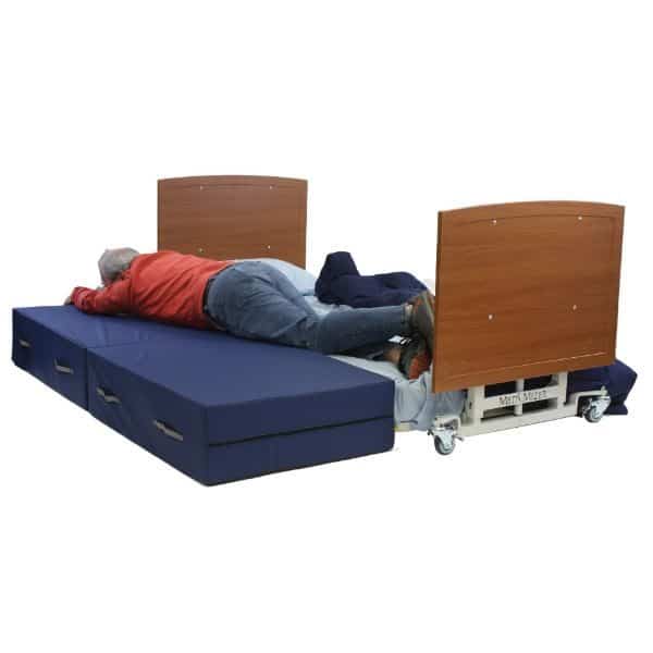 A person in a red jacket lies on a blue foldable mattress placed on a hospital bed frame with wooden head and footboards. The bed frame is metallic and has wheels, visible against a plain background.