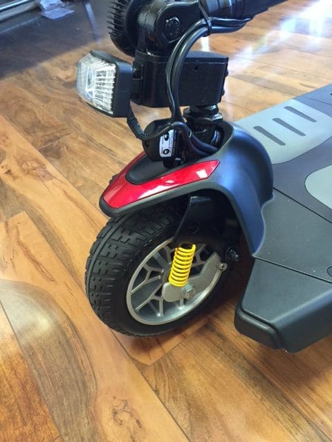Close-up of a scooters front wheel with a yellow suspension spring, black tire tread, red fender, and silver rim. The background shows a shiny wooden floor.