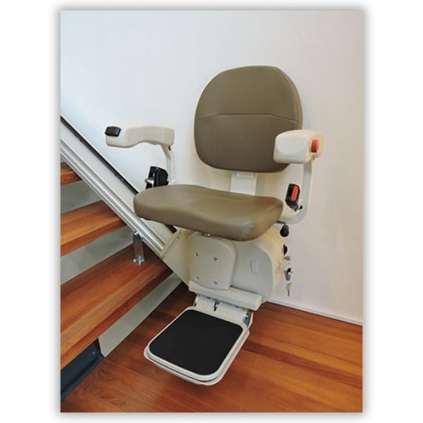 A stairlift is installed beside a wooden staircase. It features a padded seat, armrests, and a footrest, designed for mobility assistance in ascending and descending stairs safely.
