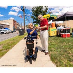 An elderly woman riding a Go-Go Elite Traveller 3 Wheel Mobility Scooter receives a drink from a smiling elderly man. Theyre near a lemonade stand with various beverages, surrounded by buildings and an American flag in the background.