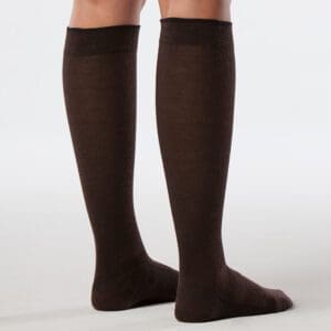A person wearing knee-high brown socks standing on a light-colored floor. The socks have a black band at the top. The background is plain and neutral.