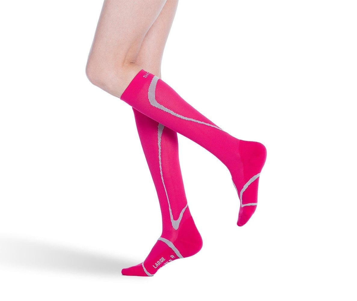 A person wearing bright pink knee-high compression socks with gray patterns stands on one foot against a white background. The foot is slightly raised, showcasing the design and fit of the socks.