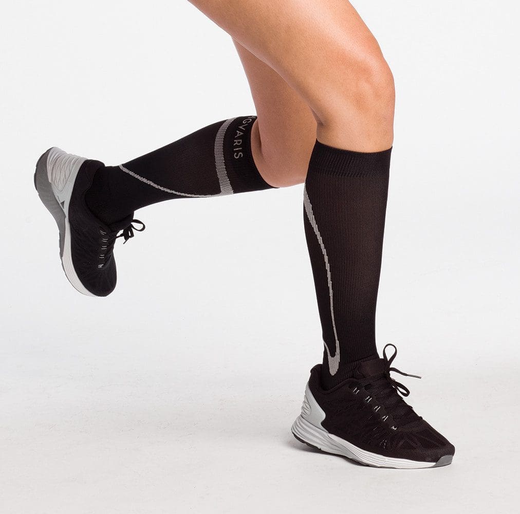 A person wearing black knee-high compression socks and black athletic shoes with gray accents. The legs are slightly bent as if about to take a step or run, with a white background.