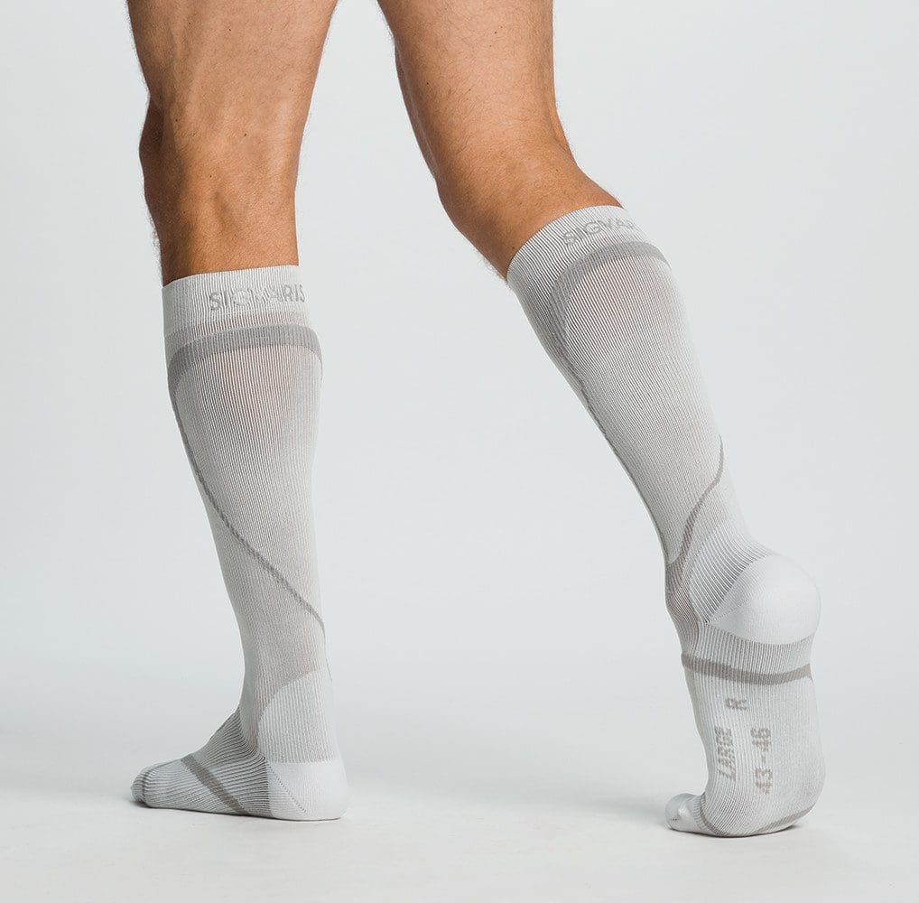 A person wearing knee-high white compression socks stands on one leg, with the other leg slightly bent and lifted behind. The background is a plain light gray.