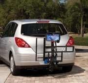 A silver hatchback car with a bicycle mounted on a rear bike rack, parked on a paved surface. Trees and grass can be seen in the background.