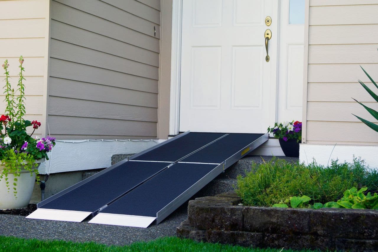 A portable wheelchair ramp is placed at the entrance of a house, leading up to the front door. The surrounding area has a well-maintained garden with green grass and colorful flowers in pots.