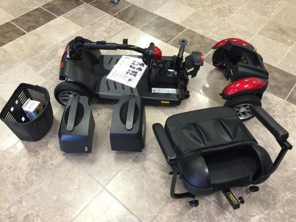 Disassembled mobility scooter with various parts laid out on a tiled floor, including a seat, two batteries, a basket, and the main base with wheels. A manual is also visible.
