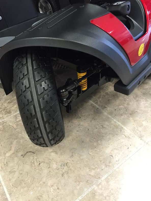 Close-up of a black and red front wheel of a mobility scooter on a tiled floor, showing the tread pattern and suspension system.