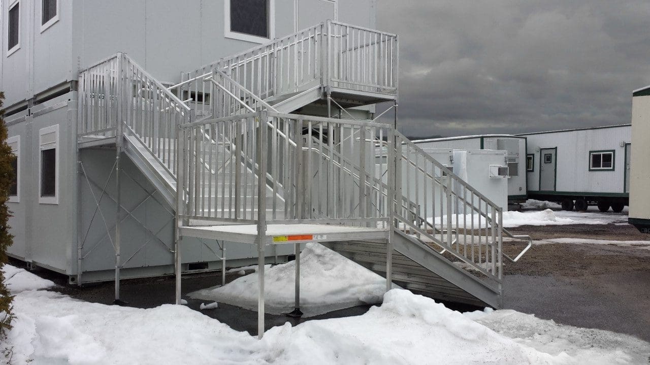 A two-story modular building with an external metal staircase. The ground is partially covered in snow, and the sky is overcast. Other similar structures are visible in the background.