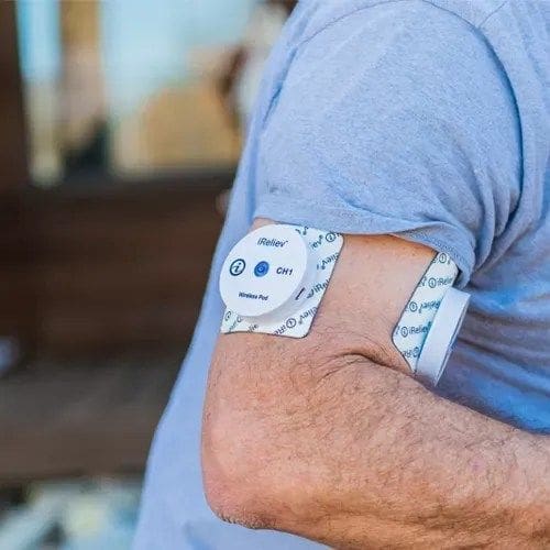 A person wearing a blue shirt uses a wireless TENS unit on their upper arm for pain relief. The white device is secured with a branded adhesive pad.