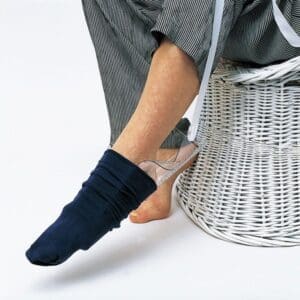 A person uses a sock aid to put on a navy sock. The sock aid is made of a flexible plastic sheet attached to two long handles. The person is sitting on a white wicker chair, wearing striped pants. The sock is partially on the foot.