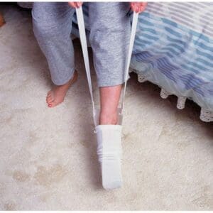 A person sitting on a bed uses a sock aid to put on a white sock. The device has long handles, and the person is wearing gray pants. The floor is carpeted.