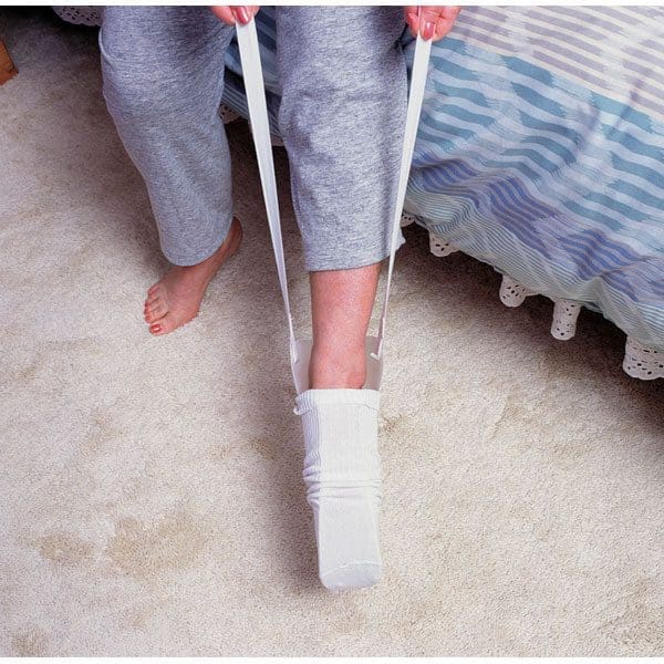 A person sitting on a bed uses a sock aid to put on a white sock. The device has long handles, and the person is wearing gray pants. The floor is carpeted.