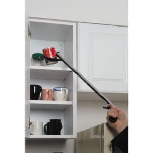 A hand uses a grabber tool to reach a can on the top shelf of a white kitchen cabinet. The shelf also holds various mugs. The cabinet door is partially open, and part of a refrigerator is visible below.