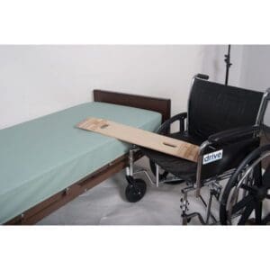 A wheelchair is positioned next to a bed with a sliding transfer board bridging the gap between them. The board has handles for easy grip. The bed has a light blue mattress cover.