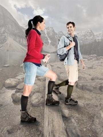 A man and woman dressed in hiking gear are standing on a wooden plank by a rocky lakeside. They are smiling and looking at each other, with misty mountains and a cloudy sky in the background.