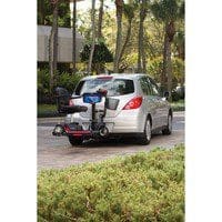 A silver car is parked on a driveway surrounded by greenery. A mobility scooter is mounted on a lift attached to the back of the car. There are trees and a building in the background.