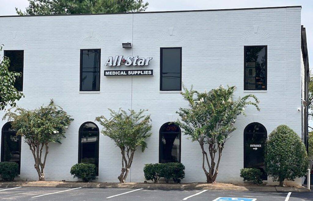 A two-story white brick building with a sign reading All Star Medical Supplies. The entrance is marked, and several bushy trees are in front. The parking lot has a space marked for handicapped parking.