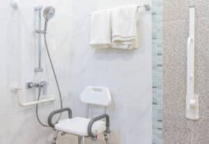 A clean, accessible bathroom with a shower featuring a hand-held showerhead, a shower chair, and grab bars. Tiled walls are partially visible, with folded white towels on a rack above.