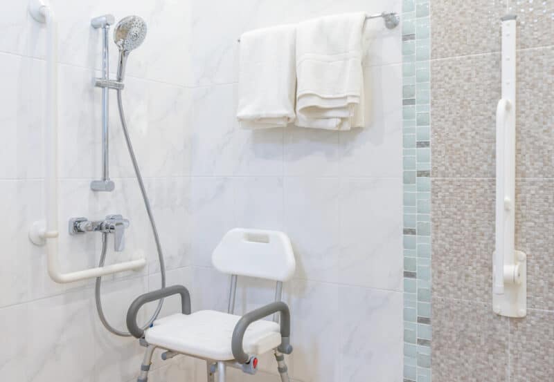 A clean, accessible bathroom with a shower featuring a hand-held showerhead, a shower chair, and grab bars. Tiled walls are partially visible, with folded white towels on a rack above.