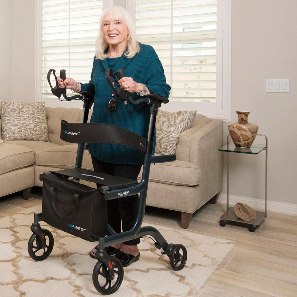 An elderly woman with white hair stands smiling in a living room using a black walker from the FAQ Walkers & Rollators collection. She wears a teal sweater and holds a pair of glasses. A couch, decorative items on a table, and large windows with blinds complete the cozy scene.