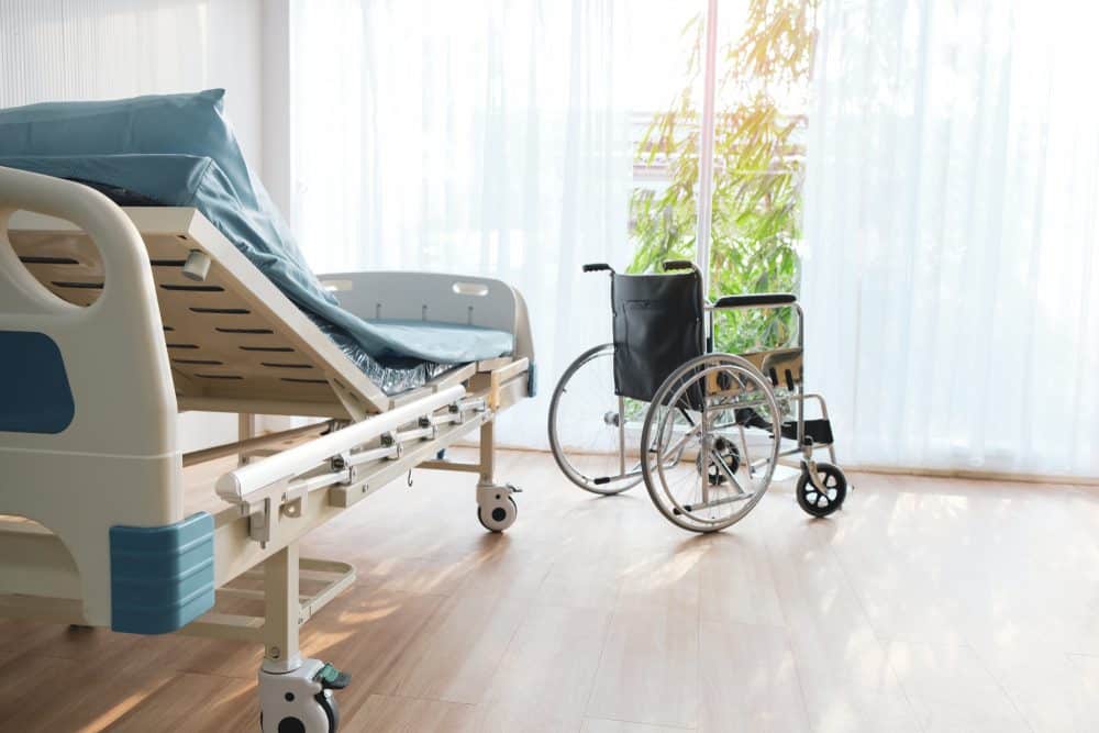 A hospital room with an adjustable bed and a wheelchair by a large window. Sunlight filters through sheer curtains, and a potted plant is visible in the background. The room has wooden flooring and a calm, airy atmosphere.