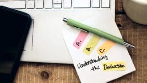 A desk with a laptop keyboard, a smartphone, and a green pen resting on a paper note. The note has colorful tabs labeled A, B, and C, with the text Understanding the Deductible nearby.