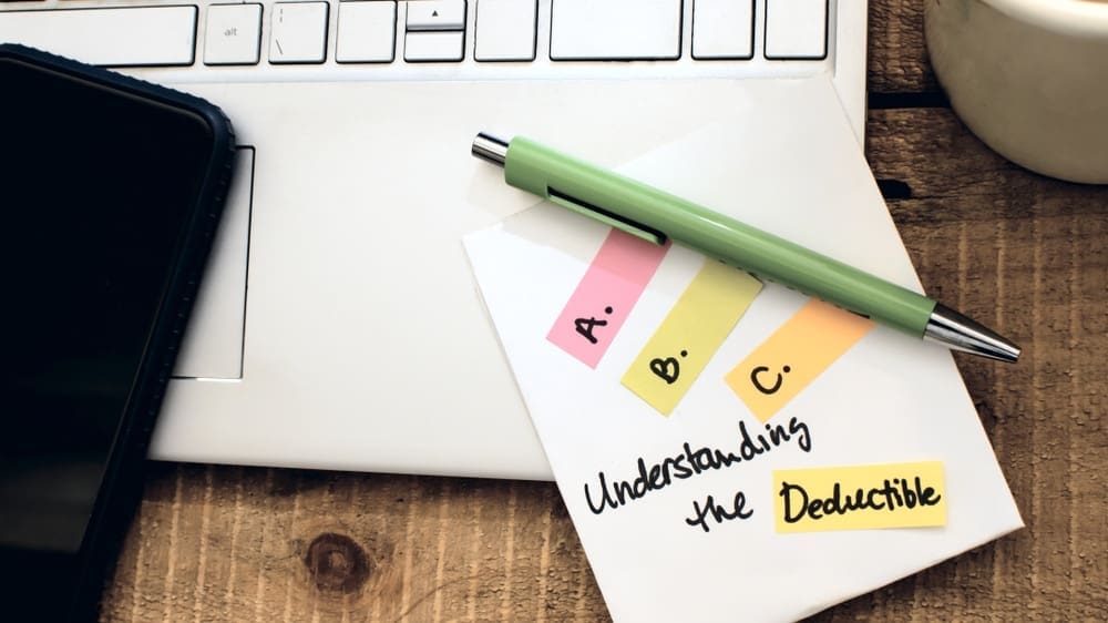 A desk with a laptop keyboard, a smartphone, and a green pen resting on a paper note. The note has colorful tabs labeled A, B, and C, with the text Understanding the Deductible nearby.