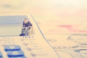 A small figurine of a person in a wheelchair is placed on a large calculator with the word TAX visible. The background is softly blurred, suggesting financial themes.