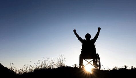 Silhouette of a person in a wheelchair with arms raised triumphantly against a sunset sky. The sun is setting on the horizon, casting a warm glow over the scene, with grass and plants in the foreground.