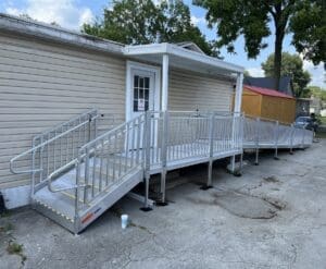 A metal wheelchair ramp with handrails enhances accessibility, leading to the entrance of a beige building. The elevated ramp features yellow safety markings and is supported by several posts. A small canopy covers the entrance, while trees and a neighboring structure are visible in the background.