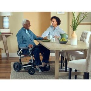 Two elderly people share a joyful moment at a dining table, with one sitting in a sleek folding power wheelchair. The room is warmly decorated with flowers and books on a side table, creating a cozy atmosphere.