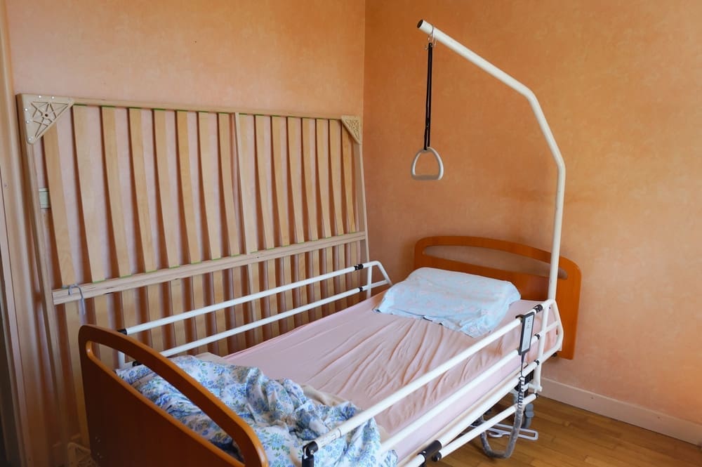 An empty hospital bed with a pink mattress, blue floral pillow, and white bed rails is positioned in a corner, illustrating the-power-of-hospital-beds-in-your-personal-space. An overhead trapeze bar is attached in the room with peach-colored walls and a wooden floor.