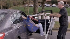A person is being transferred from a wheelchair lift into a car using a portable hoist, illustrating how exploring patient lifts can empower caregivers and enhance patient well-being. A caregiver operates the hoist with ease, the car door open against a backdrop of trees and parked cars.