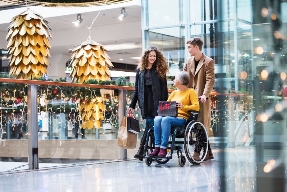 A woman in a wheelchair, accompanied by a man and a woman carrying shopping bags, is in a festive mall adorned with golden decorations and lights. They appear cheerful as they move along a shiny floor, suggesting holiday shopping.