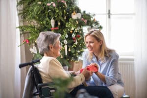 A young woman hands a small, red polka-dot gift to an older woman in a wheelchair. They are smiling warmly at each other in front of a decorated Christmas tree adorned with lights and ornaments.