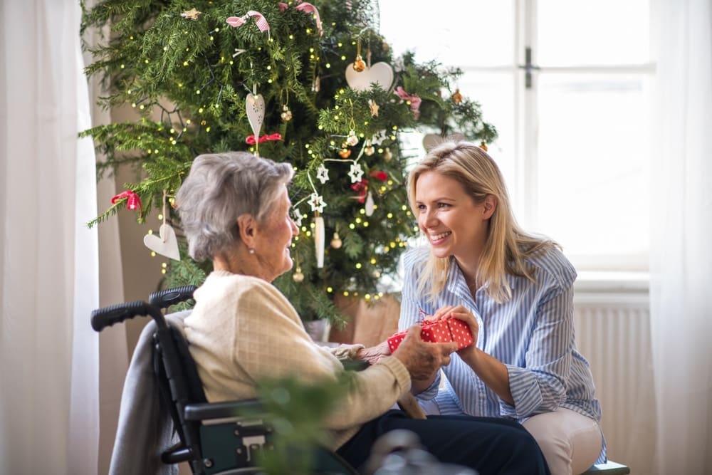 A young woman hands a small, red polka-dot gift to an older woman in a wheelchair. They are smiling warmly at each other in front of a decorated Christmas tree adorned with lights and ornaments.