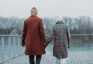 Braving the chill, two people walk hand in hand on a boardwalk beside the water, wrapped warmly in winter coats and hats. The trees stand tall against the backdrop of a cloudy sky, creating a serene setting perfect for those managing pain during cold winter months.