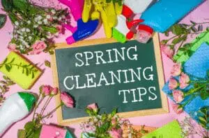 A small chalkboard with the text Spring Cleaning Tips surrounded by cleaning supplies, colorful rubber gloves, sponges, and pink roses on a pink background.