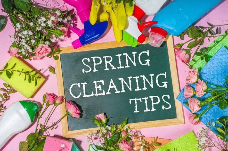 A small chalkboard with the text Spring Cleaning Tips surrounded by cleaning supplies, colorful rubber gloves, sponges, and pink roses on a pink background.